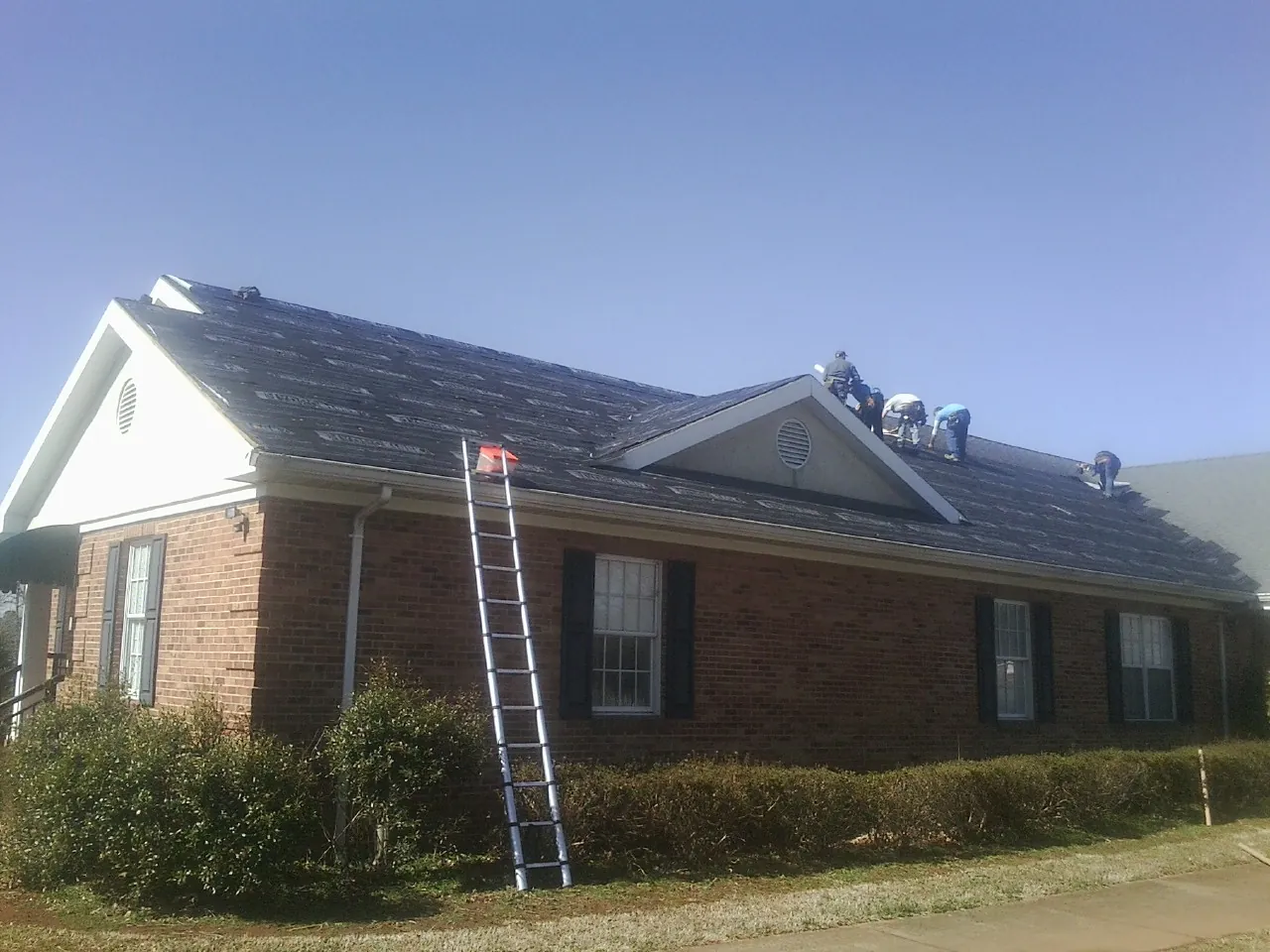 Crew tearing shingles off a brick ranch roof with a ladder set — Emergency Roof Repair work by our crew in Altmar