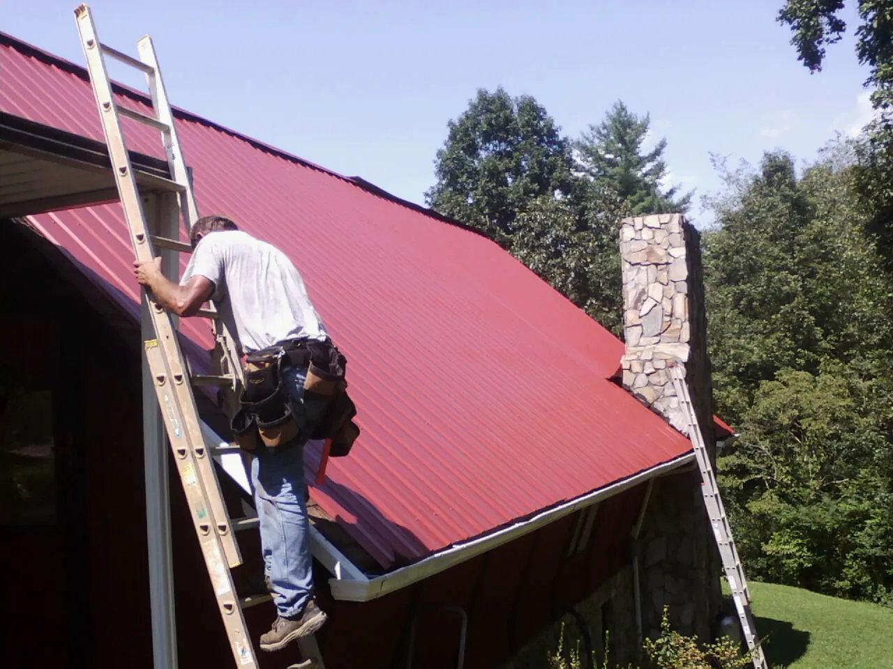 Roofer on a ladder beside a red metal roof and stone chimney — Soffit & Fascia Repair by Roofing Brothers in Altmar